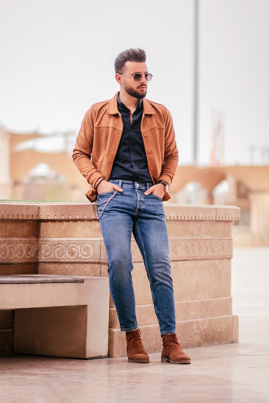 Homem adulto com jaqueta marrom e calça jeans, apoiado em uma parede de pedra clara.