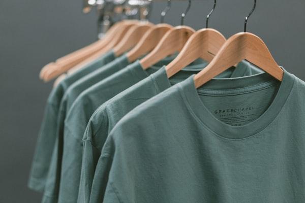 Camisetas de algodão em tom claro penduradas lado a lado em cabides de madeira.