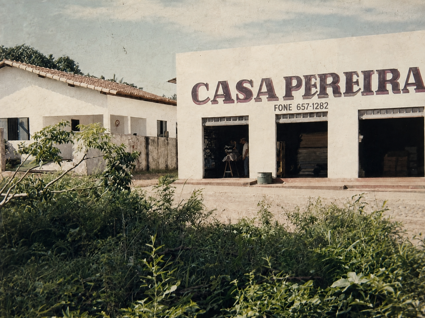 Fotografia antiga da fachada da Casa Pereira na Rua 4, com o nome da loja pintado acima das portas e vegetação em primeiro plano.
